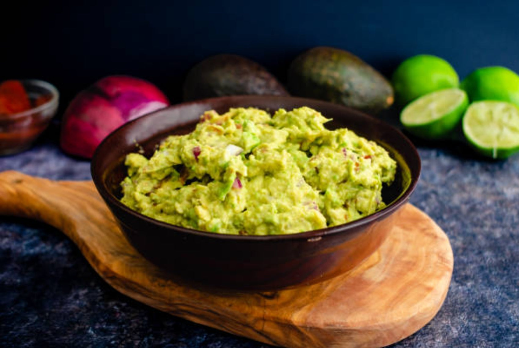 Bowl of guacamole on a wooden board with avocados and limes in the background.